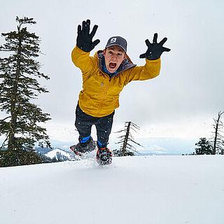 A student jumps in the snow. He is wearing a yellow jacket.